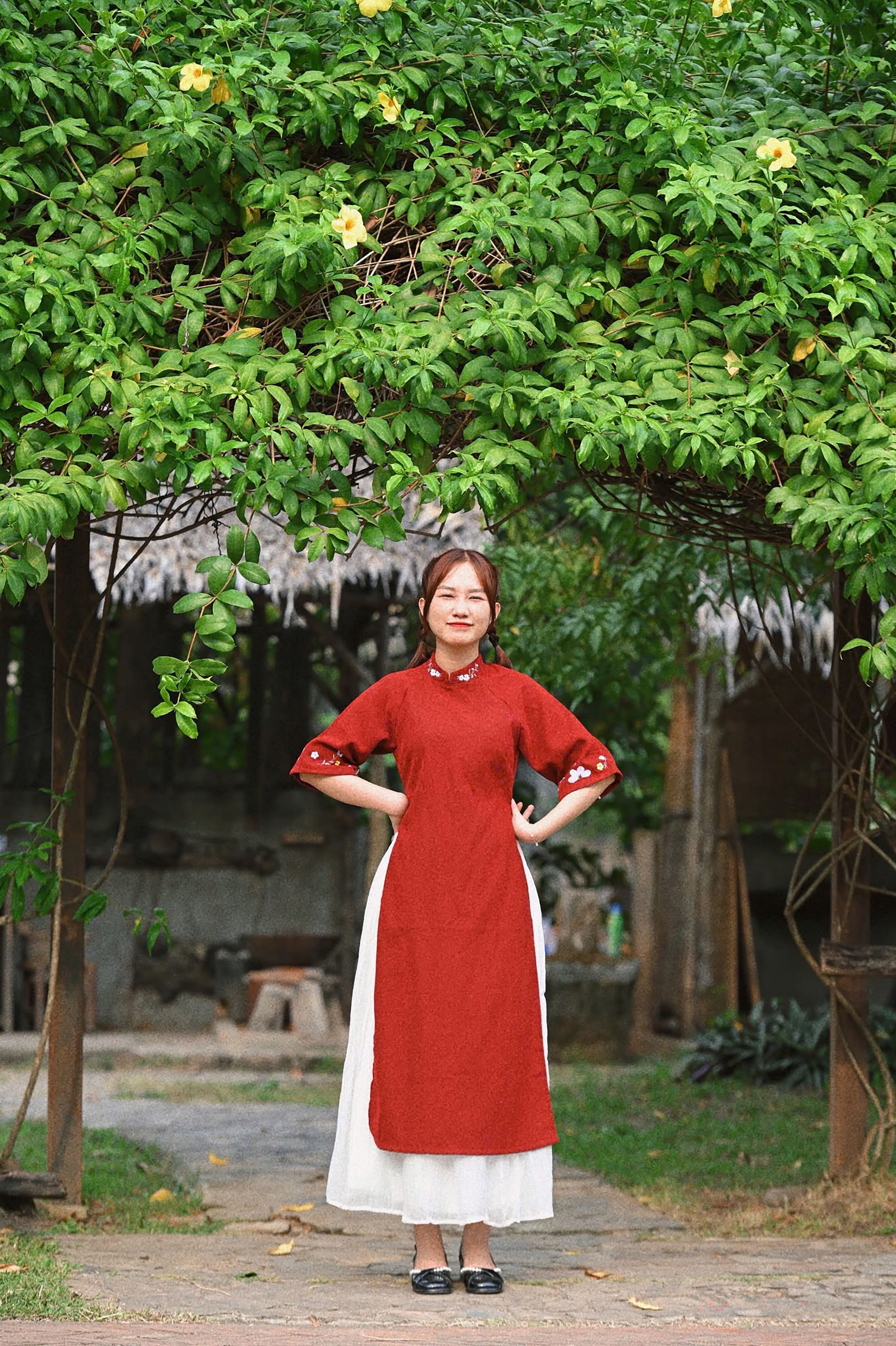 A young woman wearing a traditional red áo dài dress poses under lush green foliage in a garden.