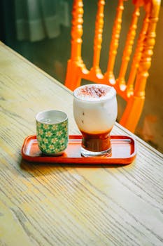 A frothy coffee and a patterned cup on a wooden table with an orange chair in the background.