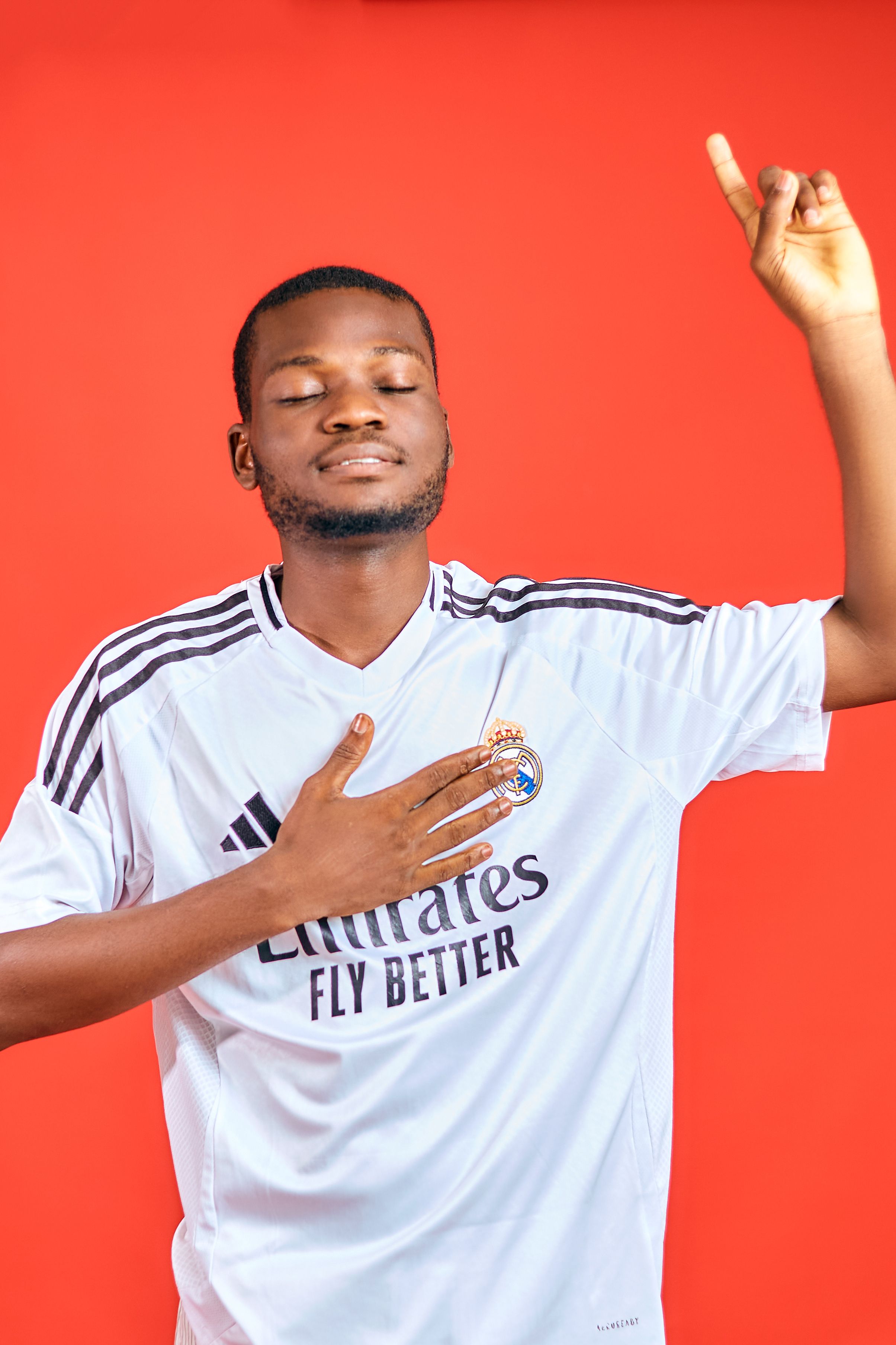 Man in white soccer jersey expresses joy with one hand on chest against red backdrop.