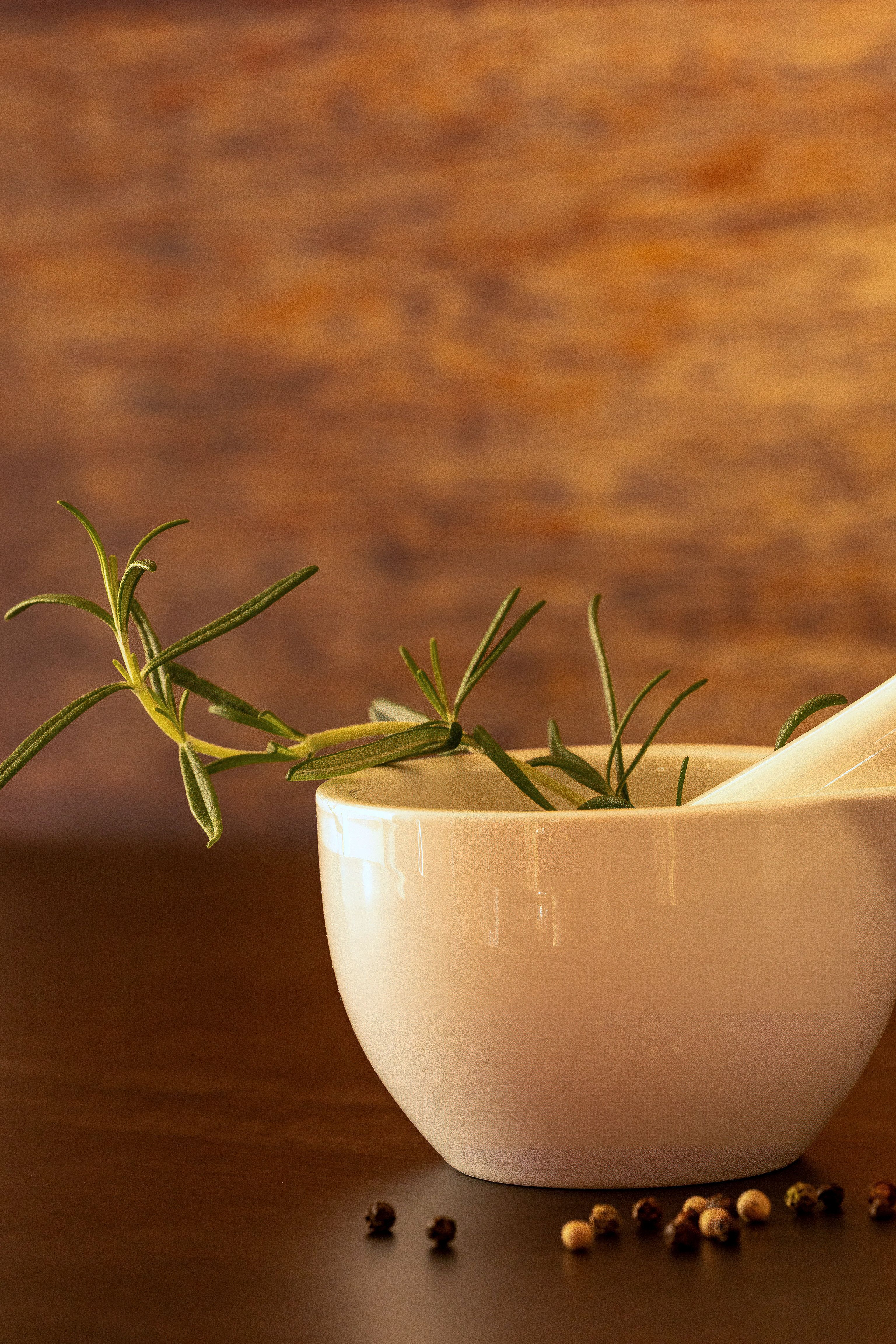 White porcelain mortar with rosemary sprig and peppercorns on wooden surface.