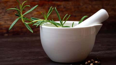 White porcelain mortar and pestle with fresh rosemary and peppercorns on a wooden background.