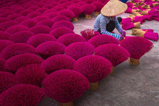 A woman in a conical hat arranges bundles of red incense sticks on the ground.