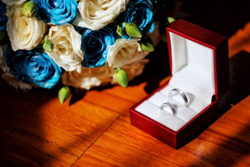 Free Stunning wedding rings in a red box with blue and white rose bouquet on wooden surface. Stock Photo
