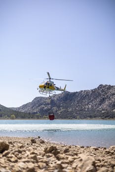 A yellow helicopter collecting water from a reservoir with mountainous backdrop.