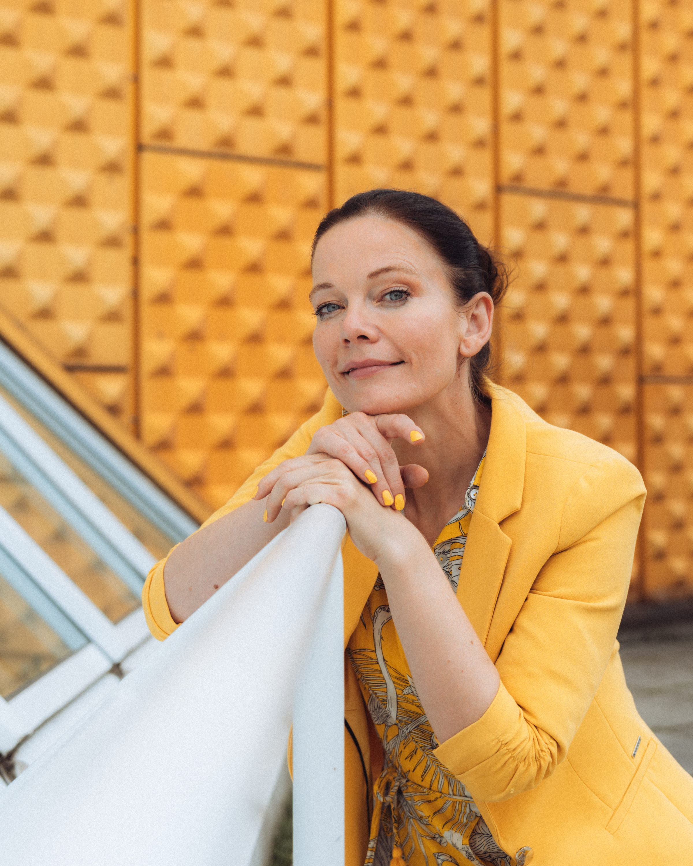 Elegant woman in a yellow blazer poses confidently against a modern architectural backdrop.
