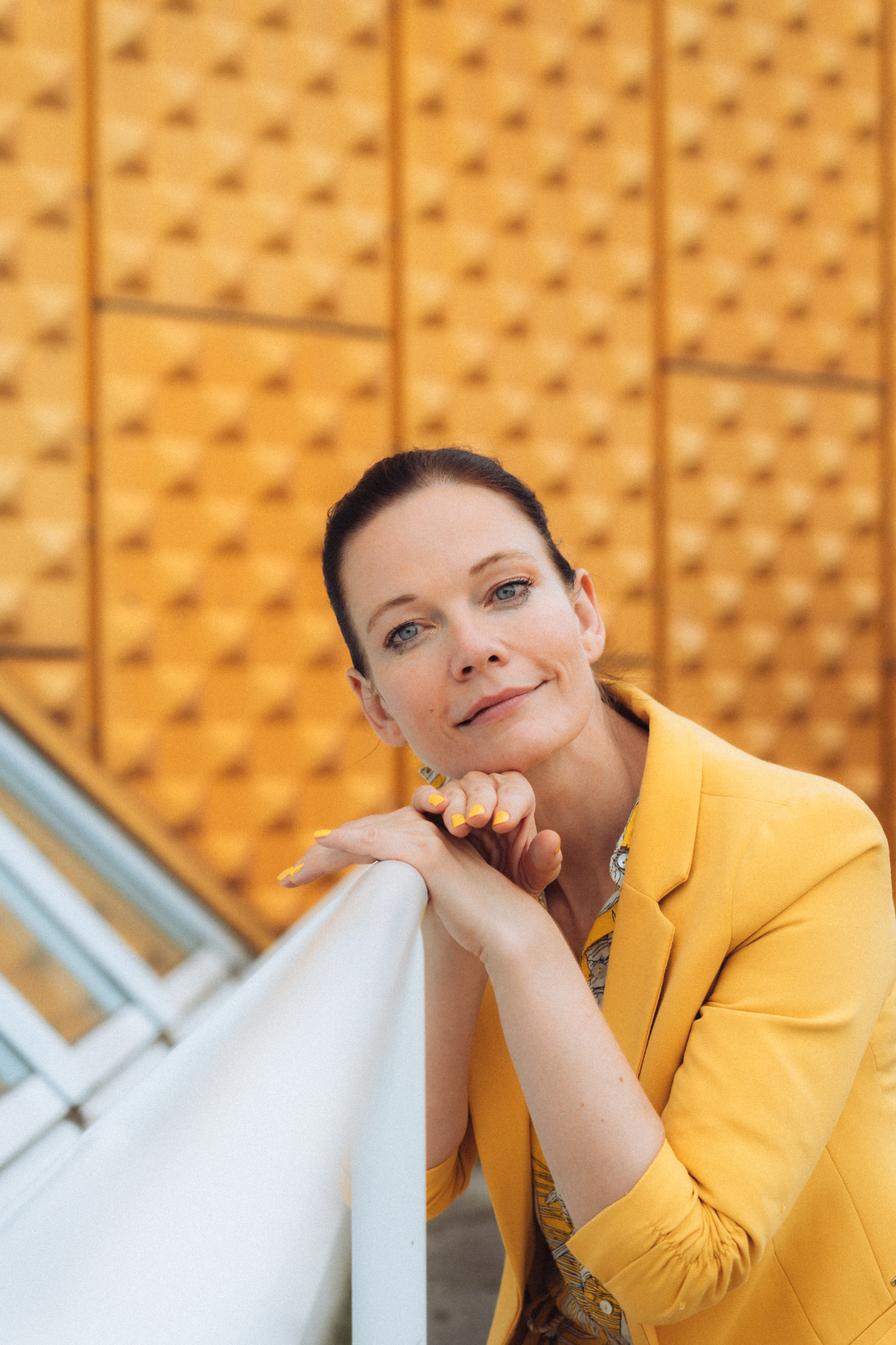 Portrait of a woman in a yellow jacket leaning on a railing against a patterned background.