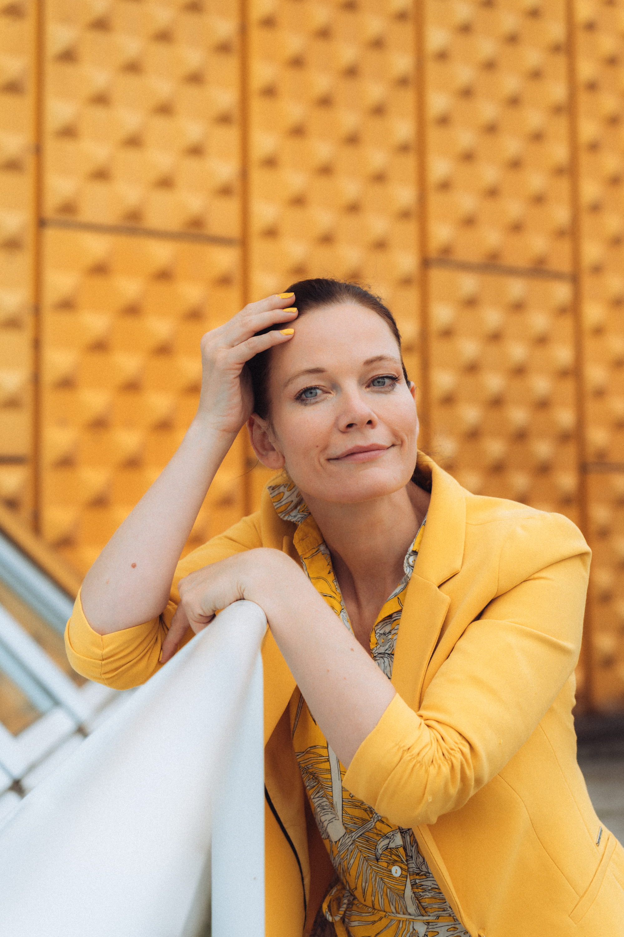 Portrait of a smiling woman in a yellow jacket against a geometric patterned wall.