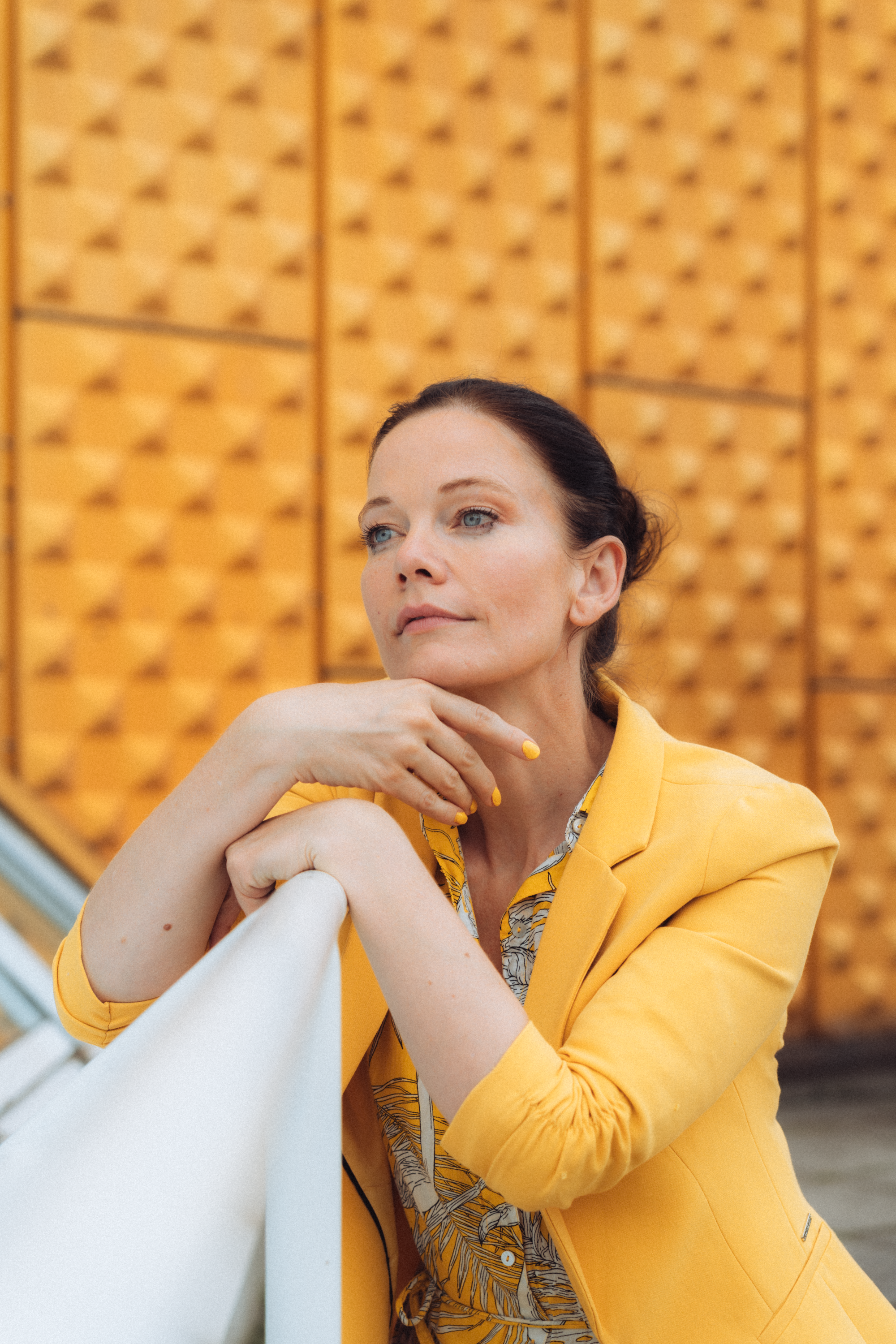 Woman in yellow blazer pondering in front of a modern geometric building.