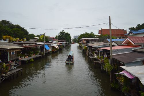 Free A river canal lined with colorful traditional Thai houses and a long-tail boat under a cloudy sky. Stock Photo