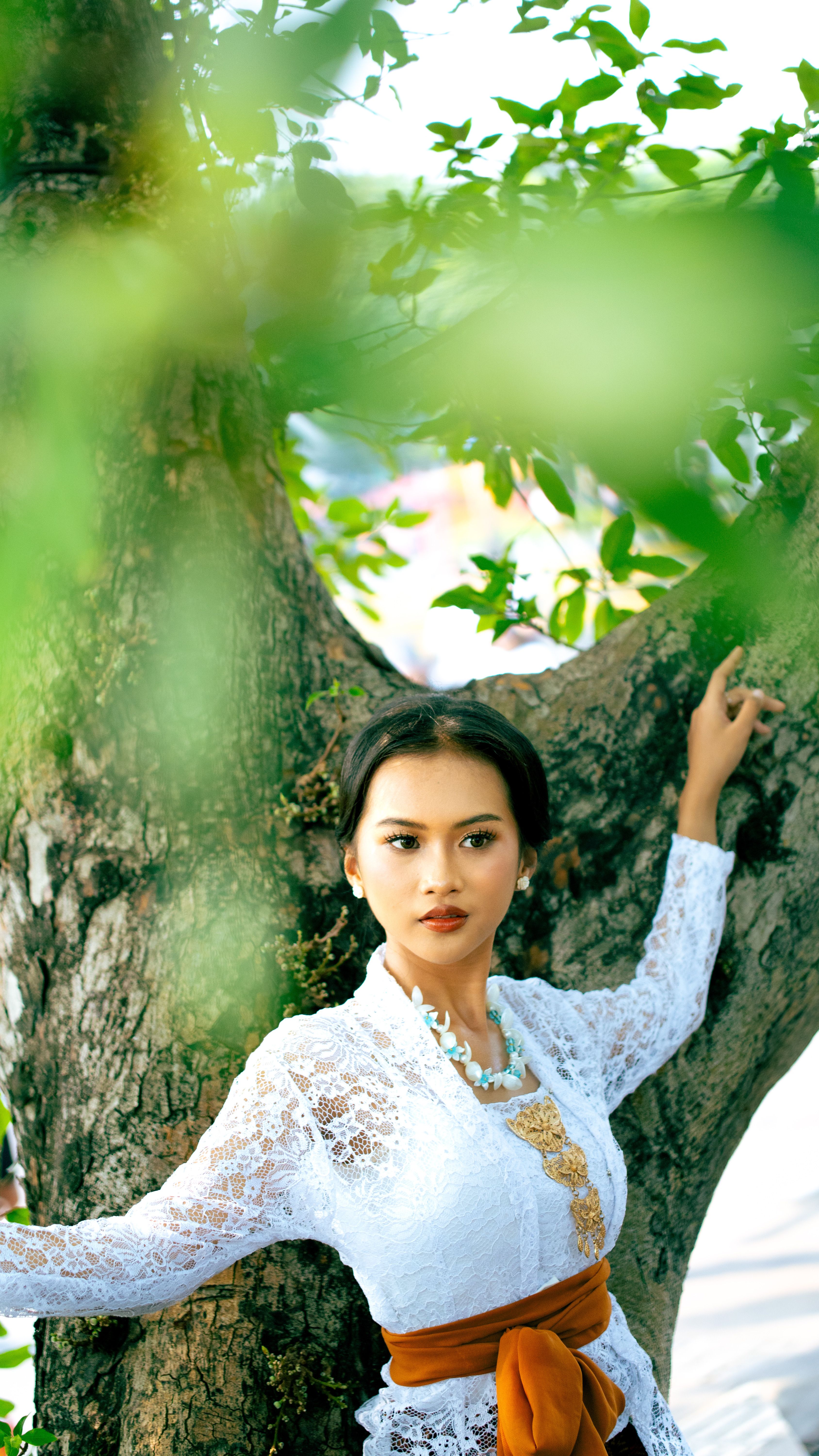 Elegant woman in white lace kebaya posing naturally outdoors by a tree in sunlight, Indonesia.