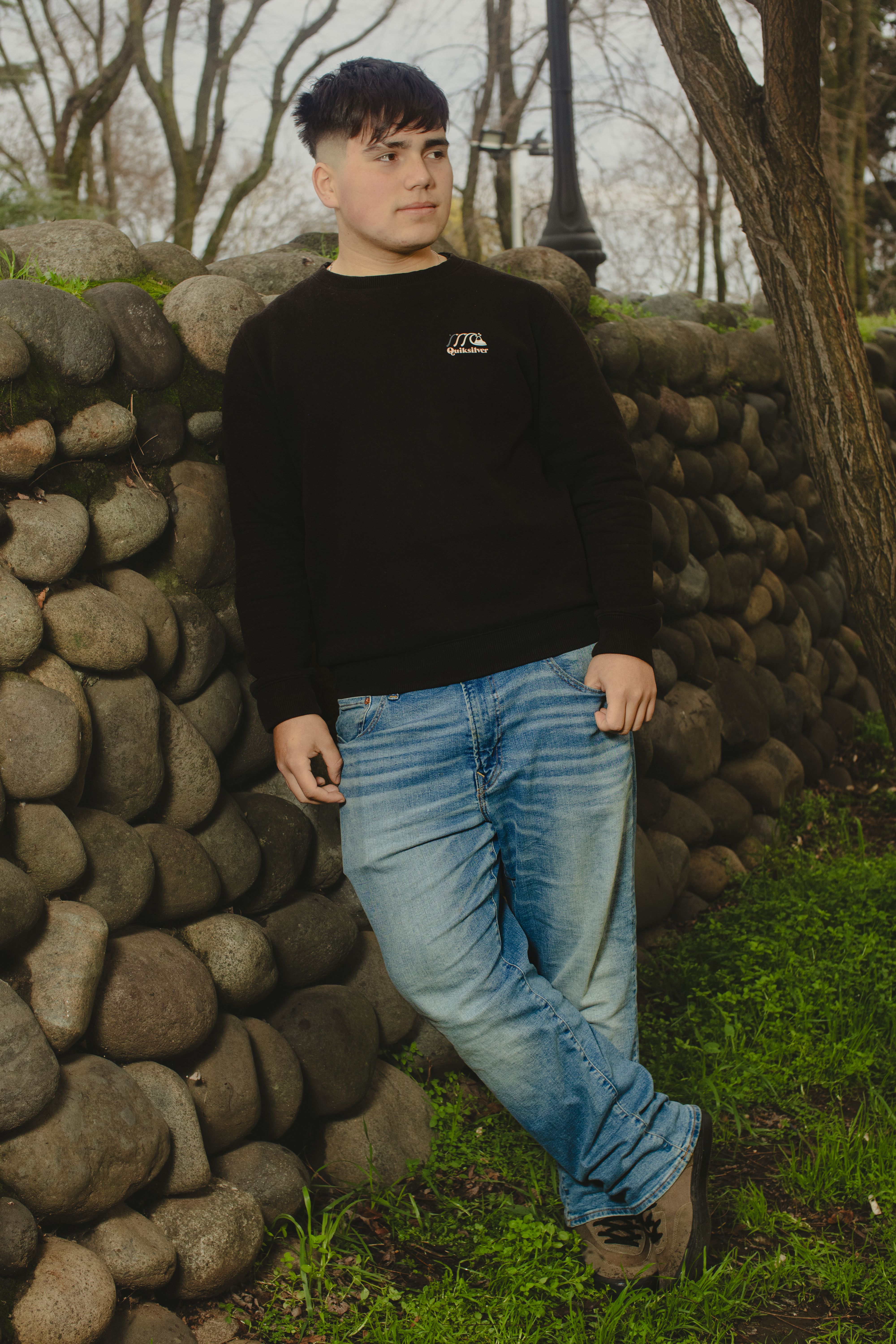 A young man in casual attire leaning against a stone wall in a park setting.