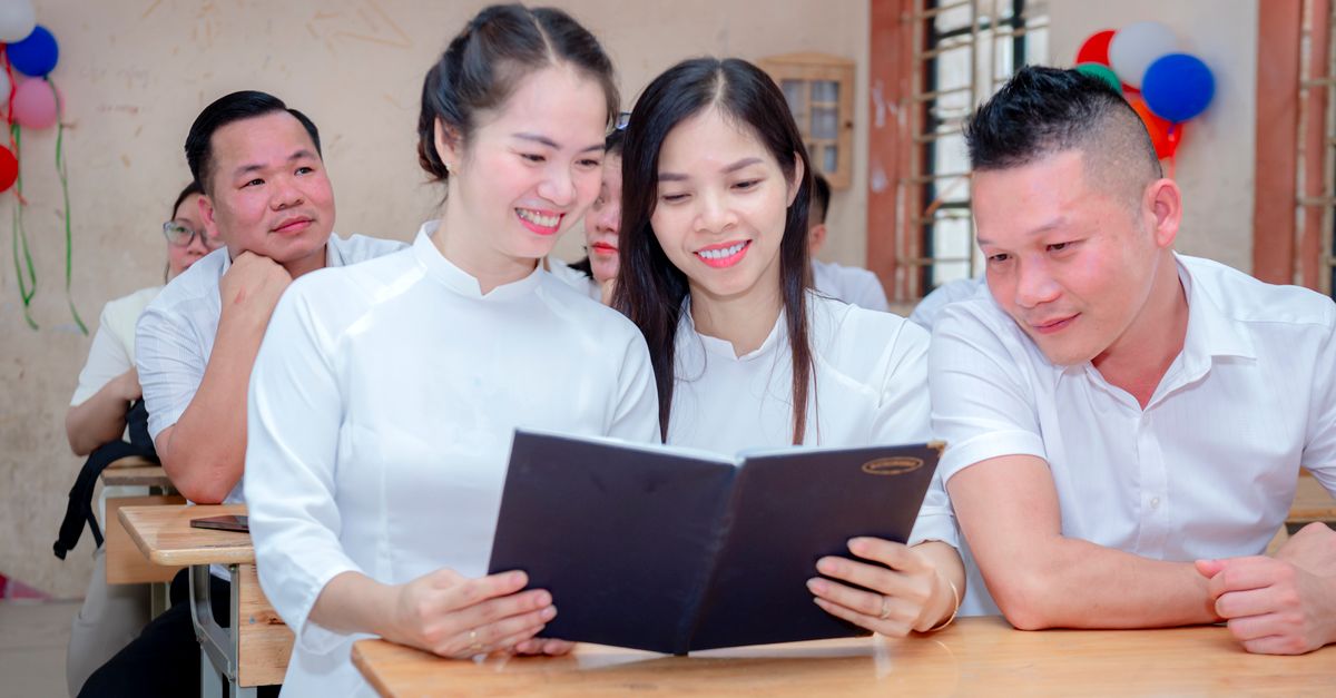 Adults gather in a classroom, smiling and engaging with a book. Positive learning environment.