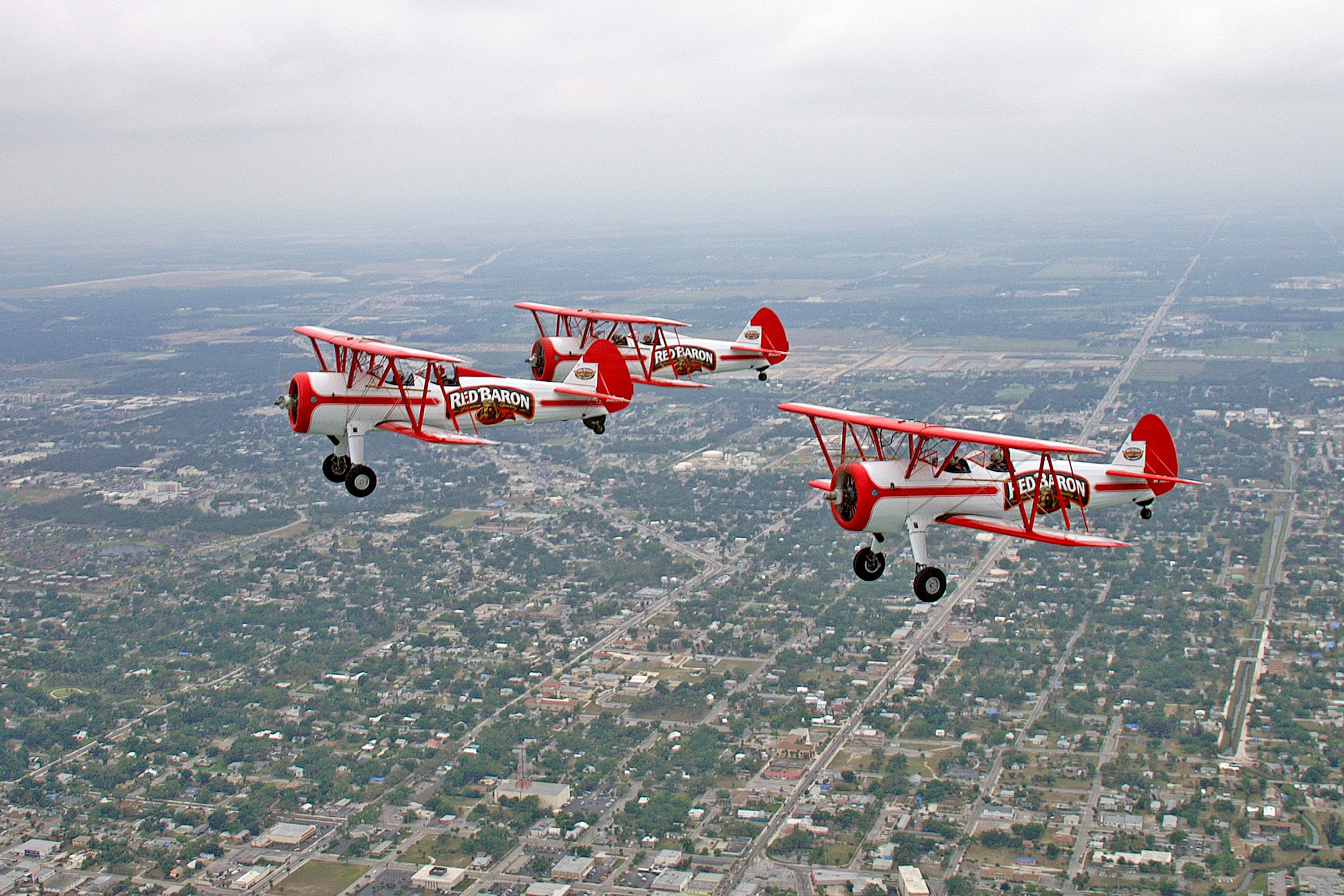 Three Red Baron biplanes flying in formation over a Florida city on a cloudy day.