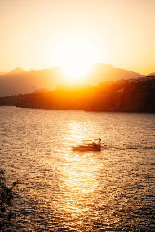Free A tranquil boat sails on the sunlit sea at sunset with mountains in the background. Stock Photo
