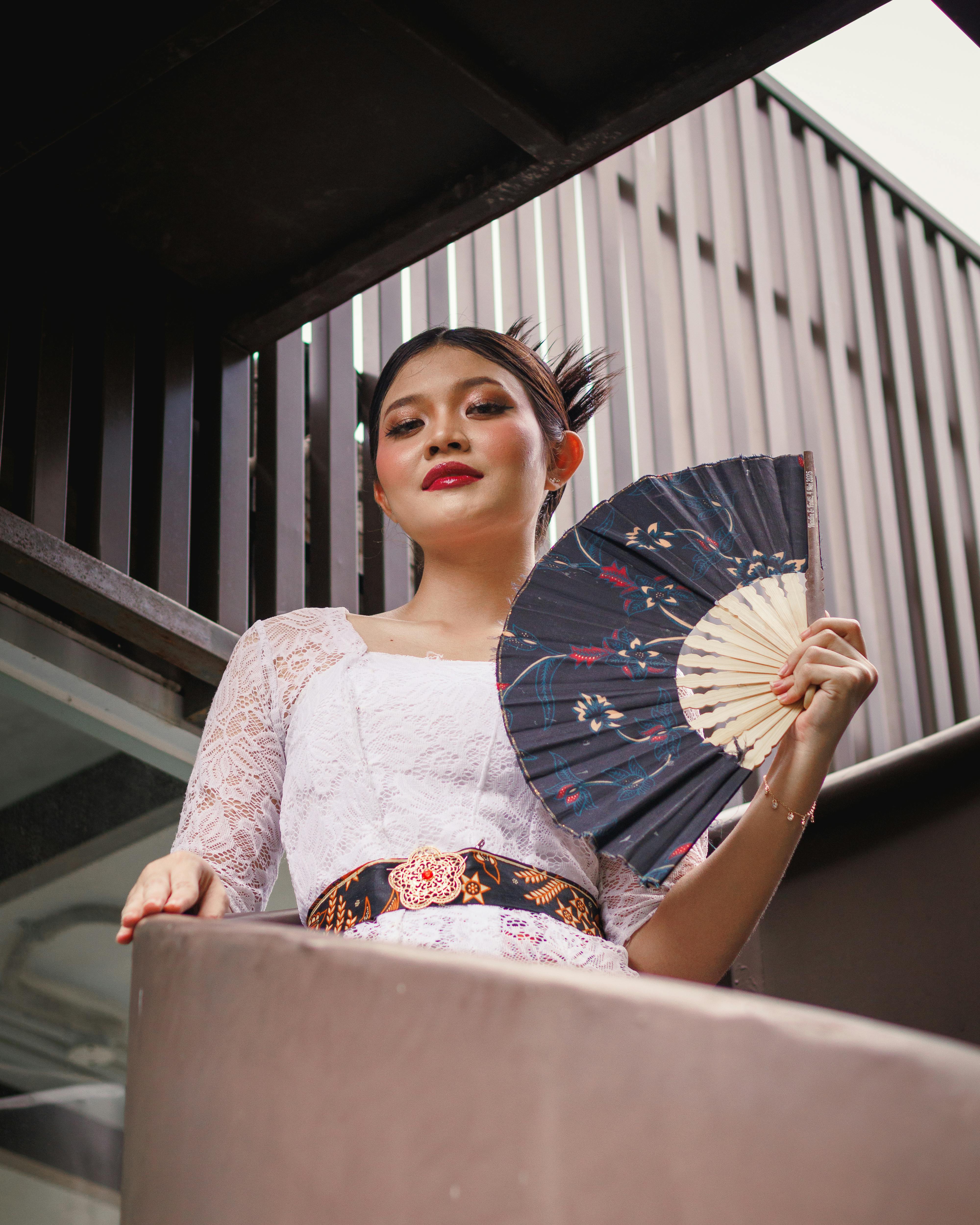 Stylish woman in traditional dress holding a fan outdoors, exuding elegance and grace.