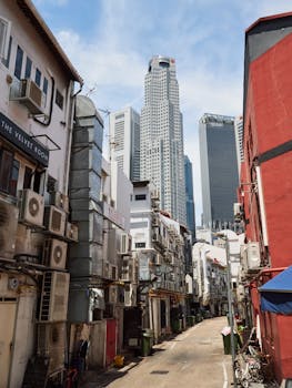 Narrow alleyway lined with buildings and air conditioners against tall skyscrapers under a clear blue sky.