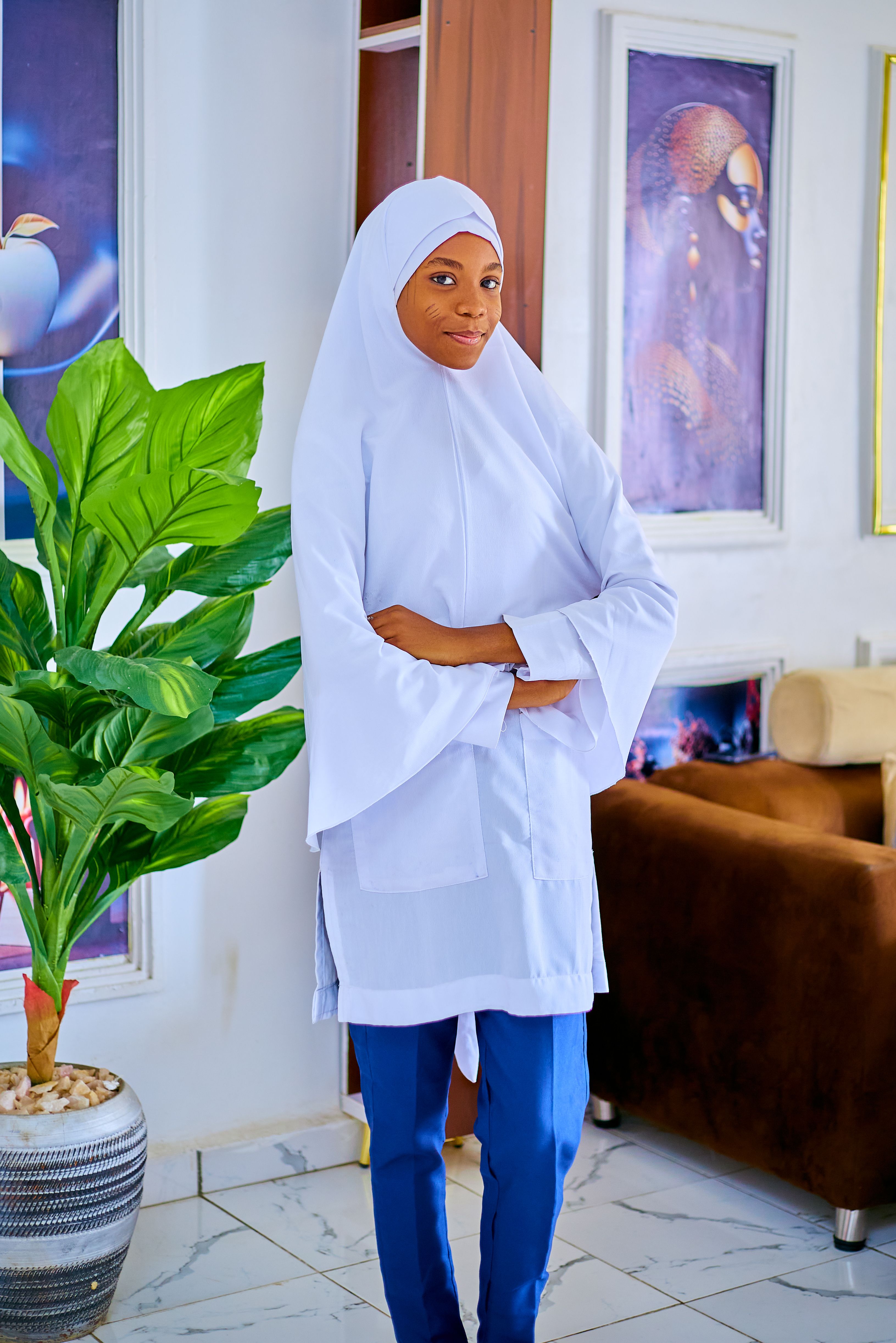 Woman in a white hijab stands confidently indoors beside a potted plant.