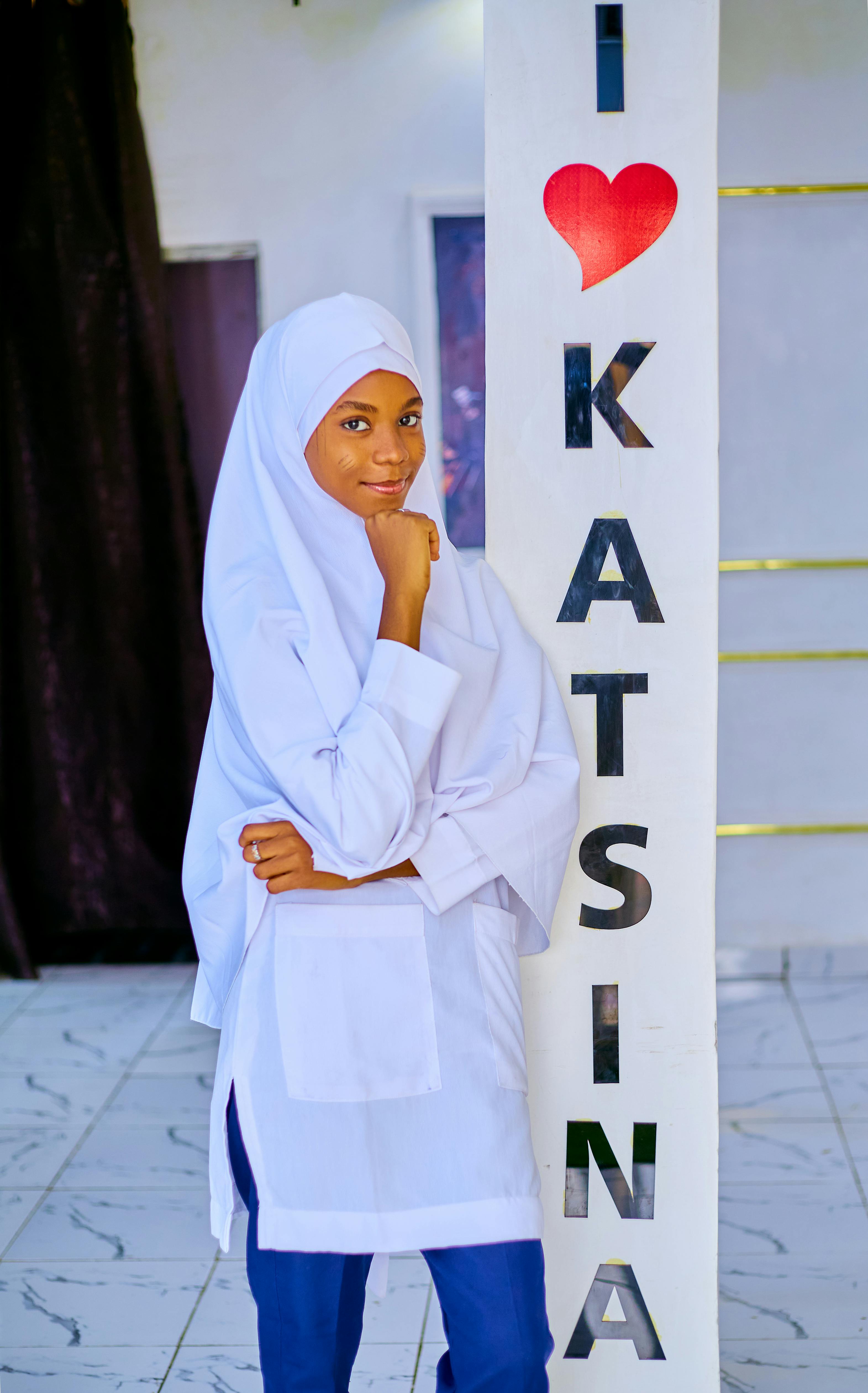 Young woman in hijab posing indoors beside 'I Love Katsina' sign.