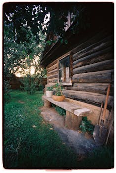 Charming log cabin exterior with garden tools and foliage at sunset, evoking a peaceful rural atmosphere.