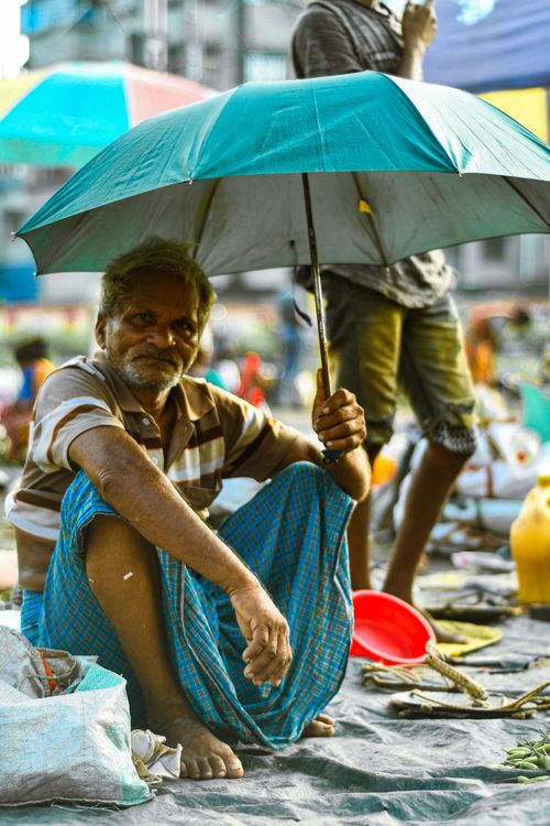 Free A street vendor sits under an umbrella protecting himself from the sun in a bustling outdoor market. Stock Photo