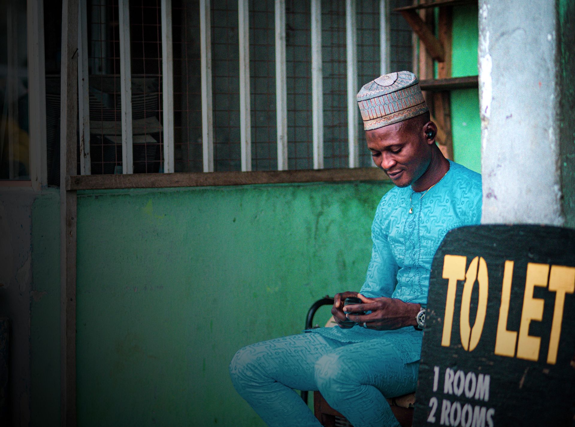 A Nigerian man in traditional attire uses his phone outside a building with a 'To Let' sign.