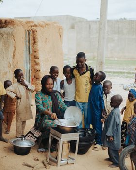 A woman cooking outdoors surrounded by children in an African village setting.