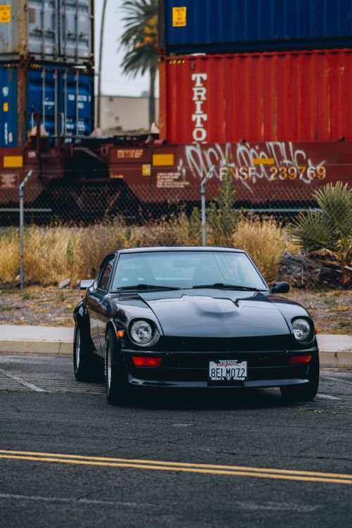 Free A vintage black car parked in front of colorful shipping containers on a sunny day. Stock Photo