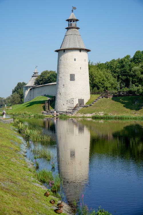 Free Historic Pskov Krom tower reflecting in the serene river with lush greenery and blue sky. Stock Photo