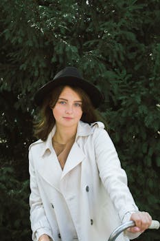 一位时尚女士头戴黑色帽子、身穿风衣，在户外郁郁葱葱的绿植前摆姿势