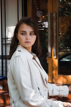 一位穿着风衣的女人站在咖啡馆外，她的肖像在自然光下显得格外优雅