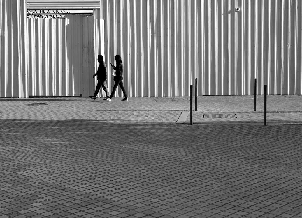Grayscale Photography of Two Person Walking Beside Wall