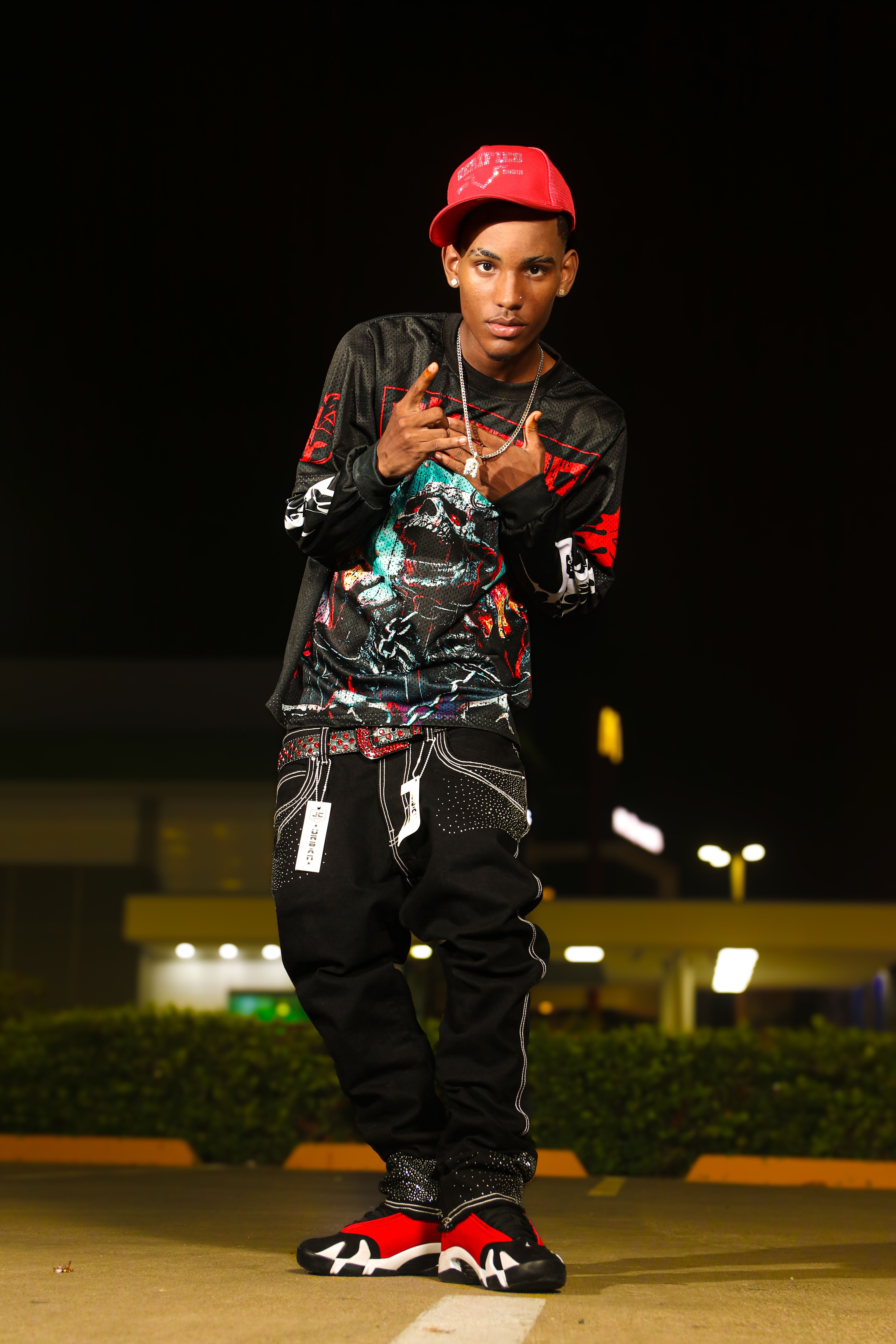 A young adult male poses in urban fashion attire at night with a vibrant setting.