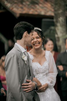 Açık havada yapılan bir düğün töreninde birbirlerine sarılan neşeli gelin ve damat gülümsüyor.