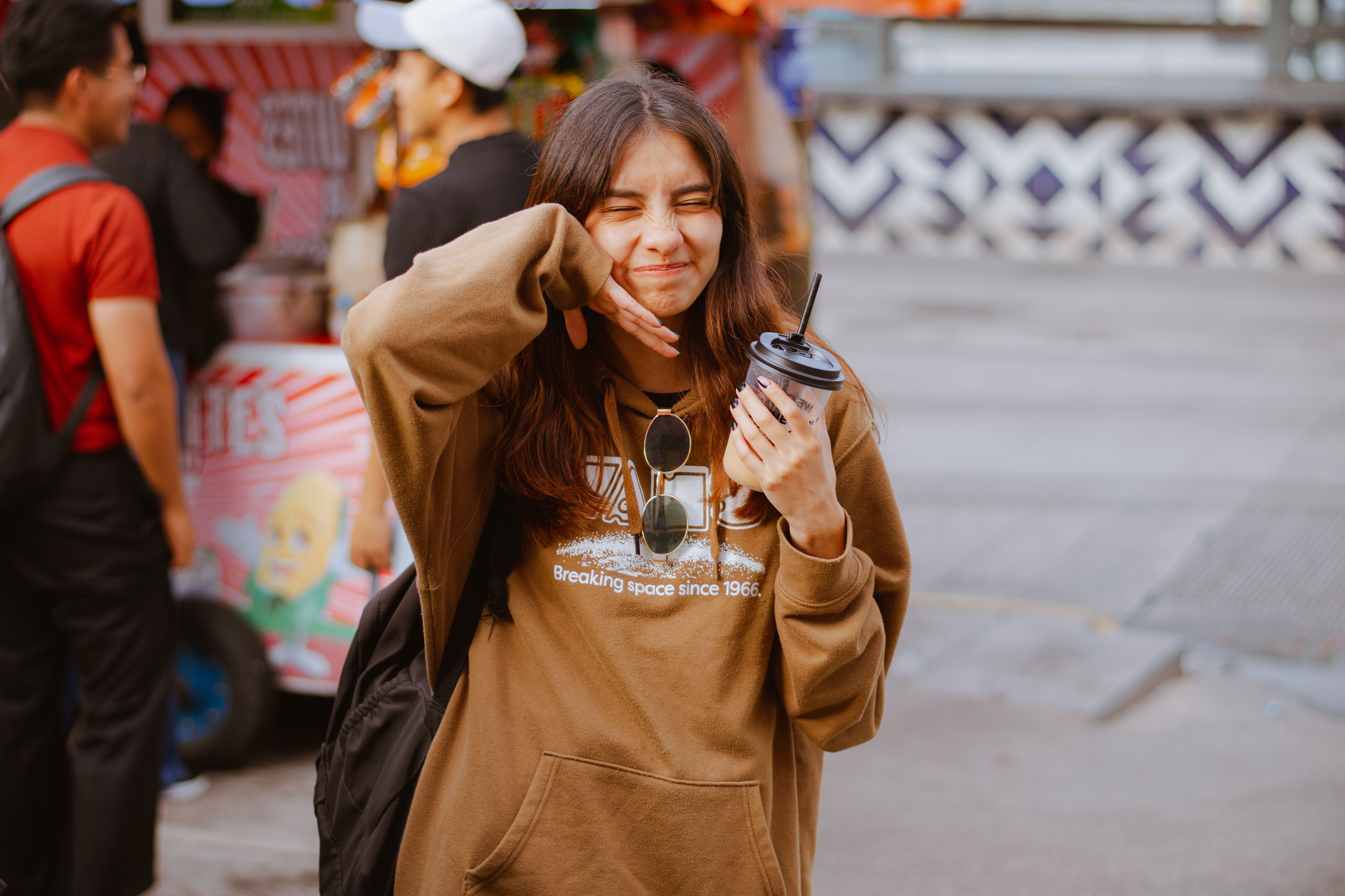 A young woman with a drink, squinting happily in a street market setting.