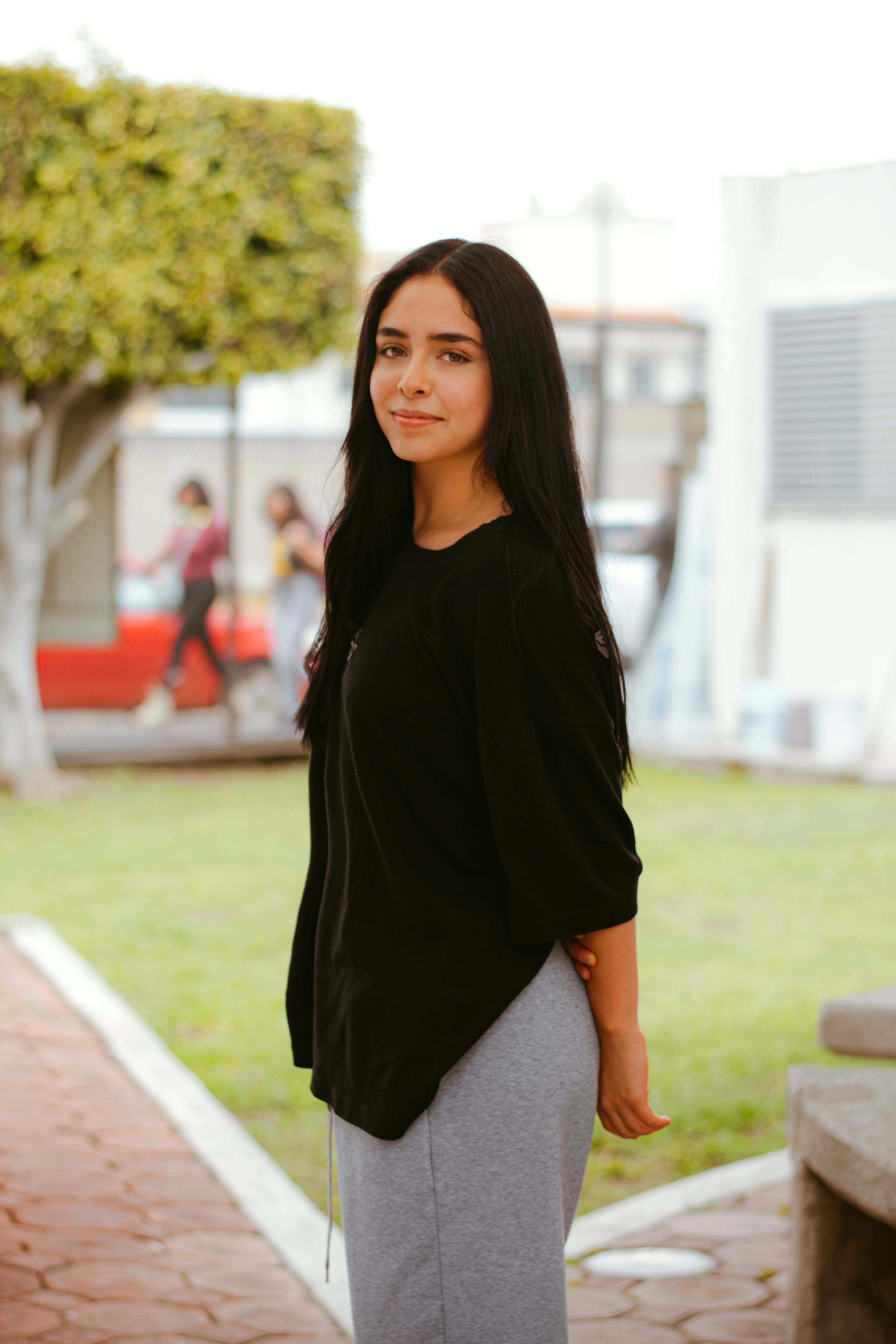 A woman stands outdoors in a relaxed pose with a soft smile on a sunny day.