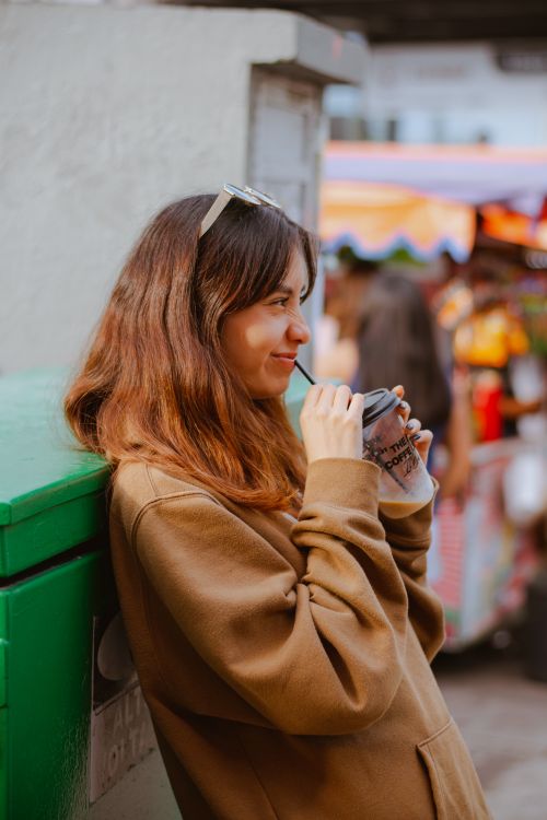 Free Young woman in a brown hoodie sipping a drink on a lively street. Stock Photo