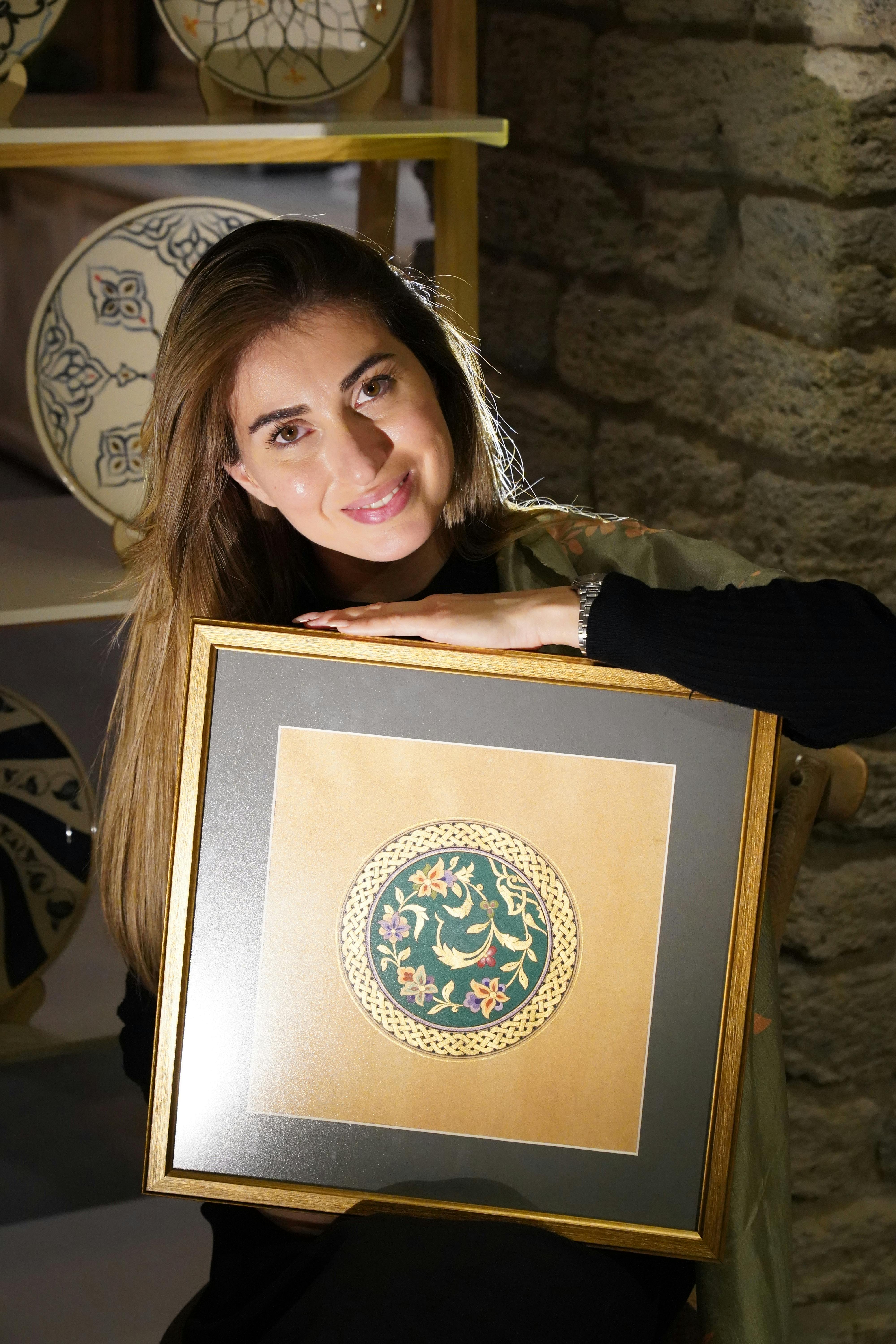 A woman proudly showcasing a framed ceramic plate with intricate floral design indoors.