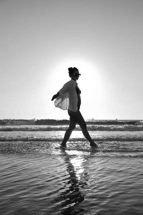 Free Silhouette of a woman walking along the beach with the sun setting behind her. Stock Photo