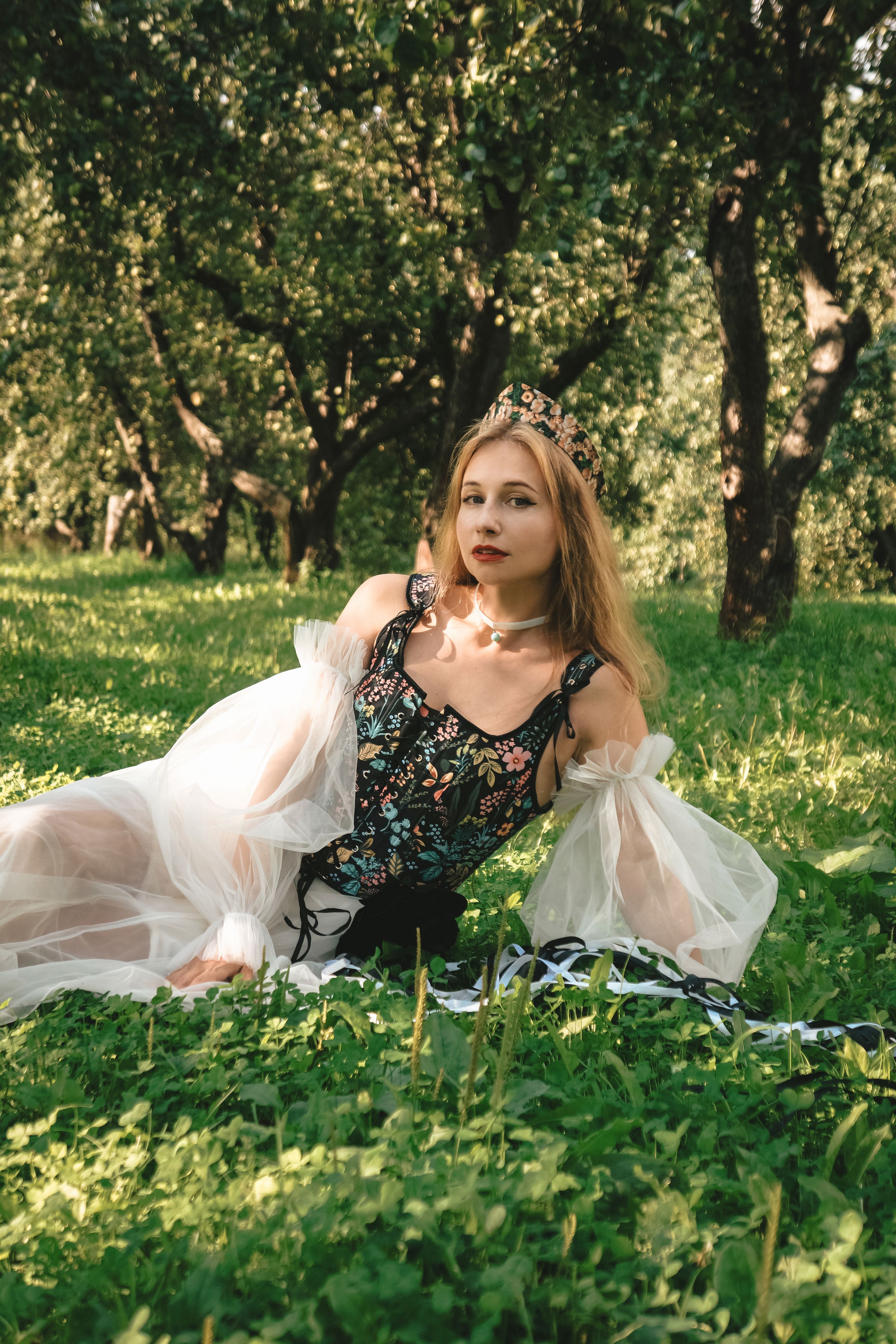 A woman in a floral dress and crown poses gracefully in a sunlit orchard setting.