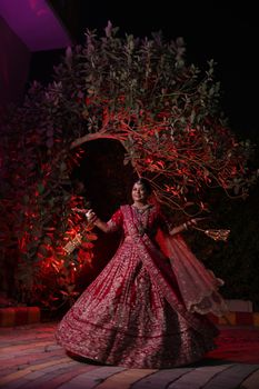 A stunning bride in a red lehenga twirls under a tree with dramatic lighting at night.