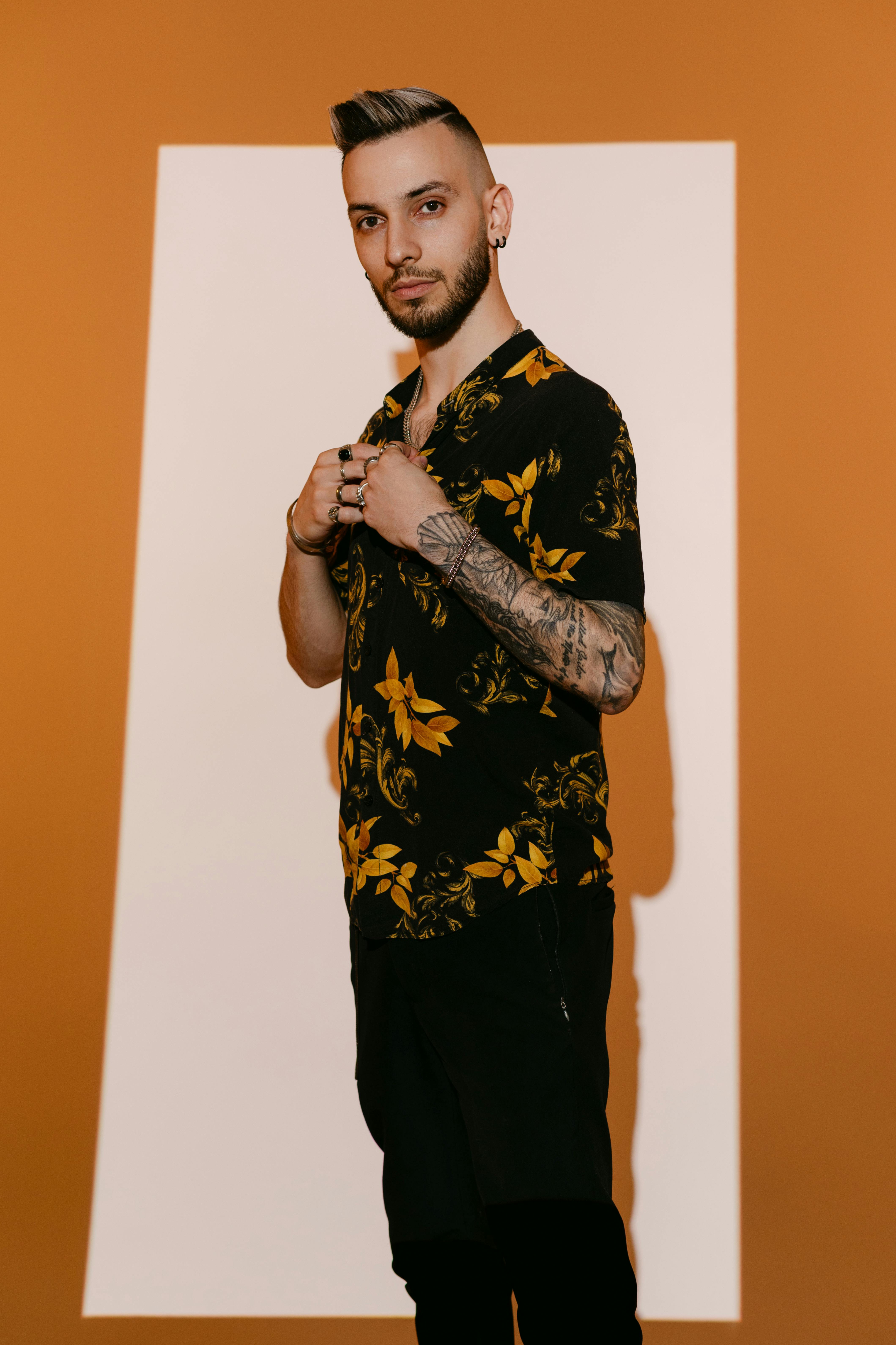 Portrait of a stylish man with tattoos wearing a floral shirt, standing confidently against an orange background.