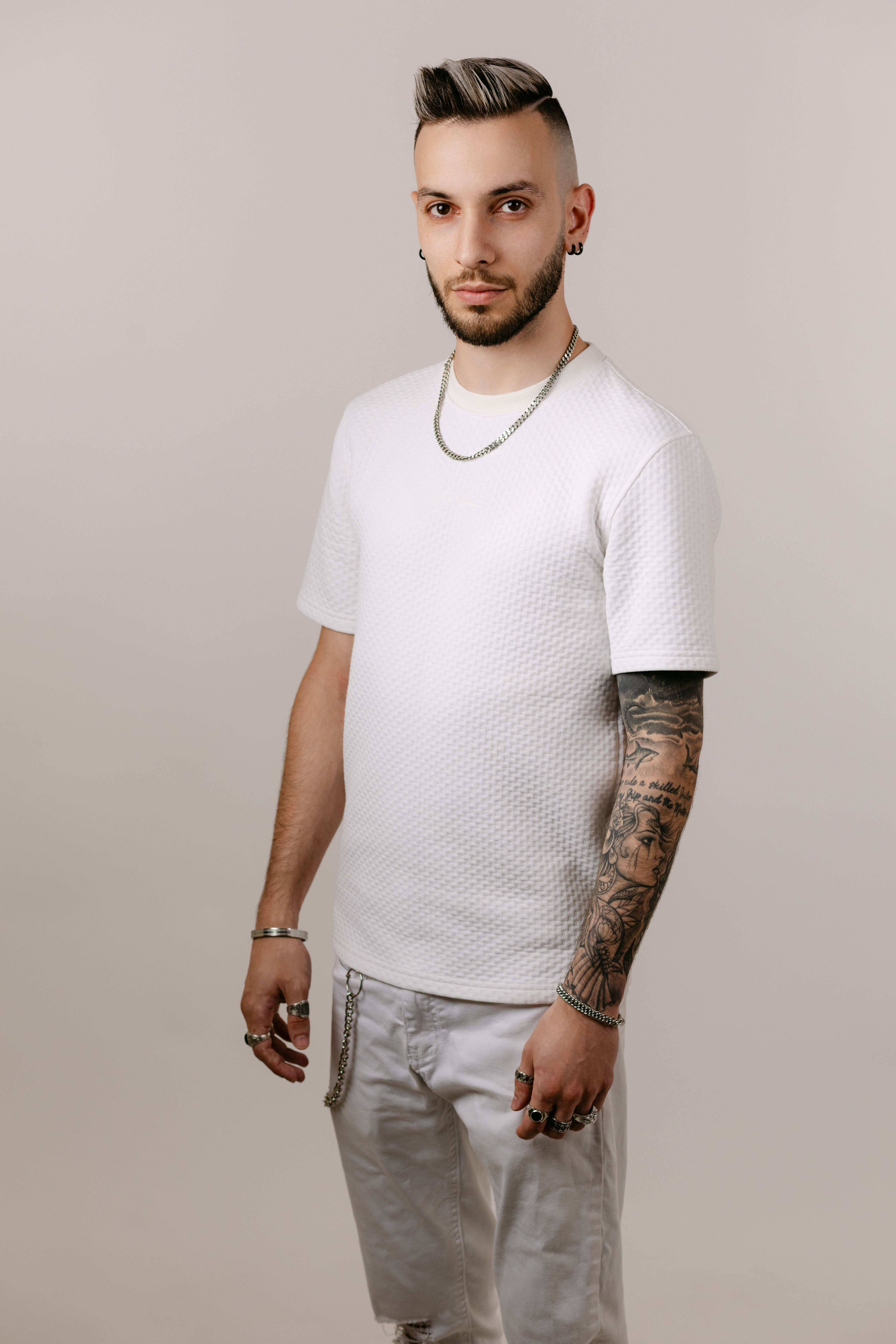 Stylish young adult male wearing white outfit and showcasing tattoos in a studio setting.