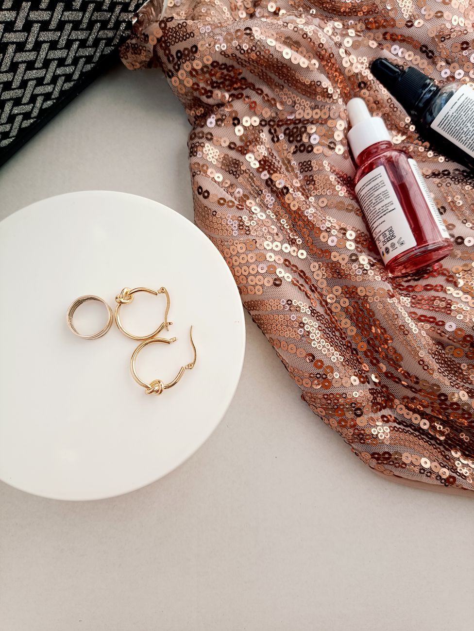 Gold jewelry on white pedestal with sequin fabric and skincare products, elegant and glamorous.