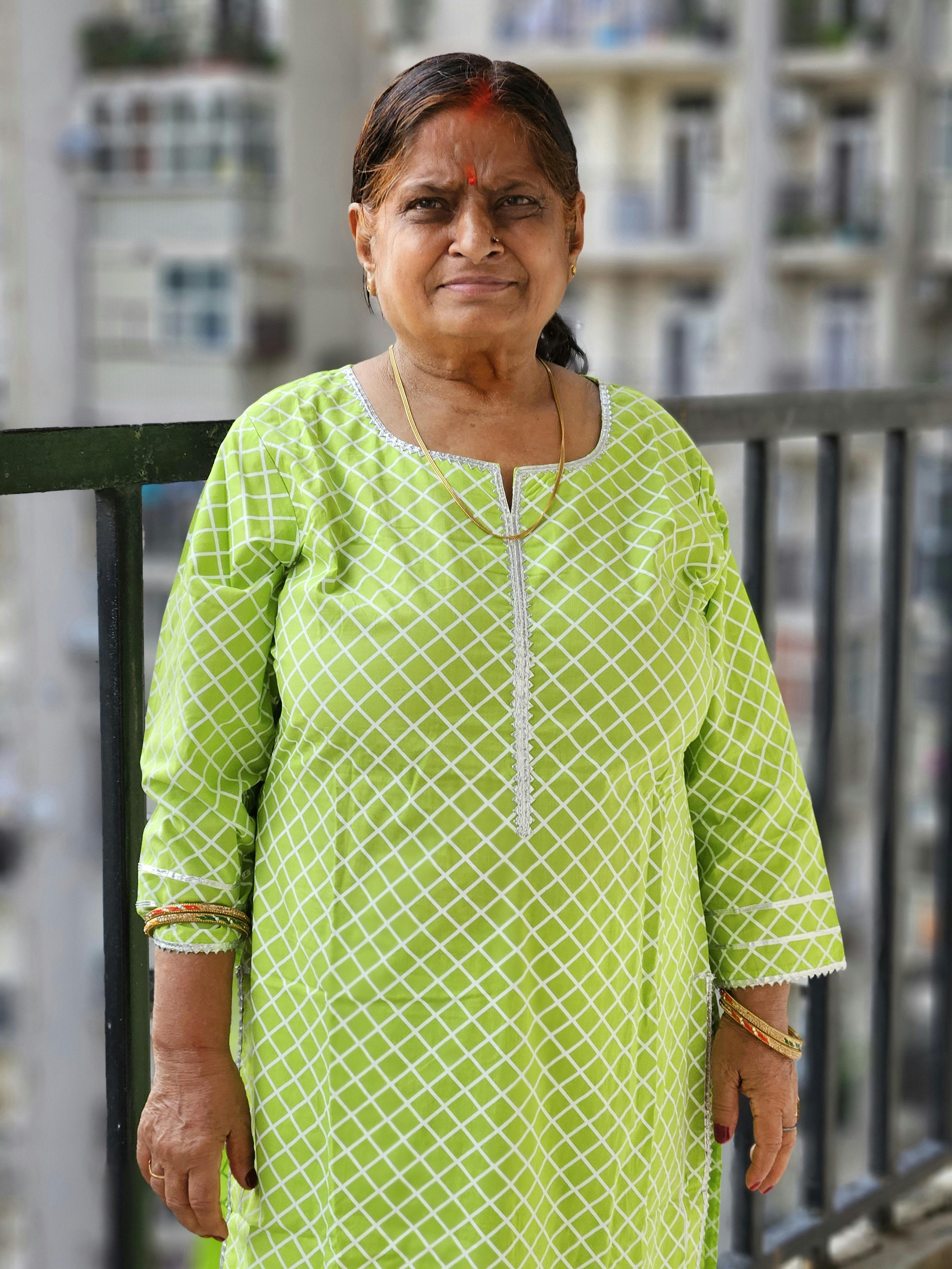 Elderly woman standing on balcony in green kurta, smiling warmly.
