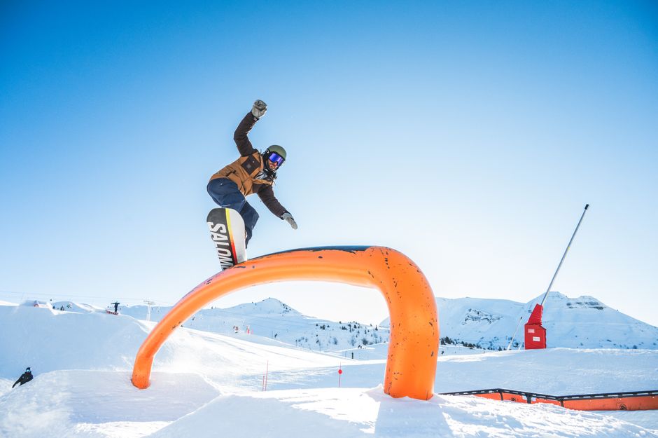 Snowboarder executing a high jump over a curved obstacle on a sunny day at a snowy resort.