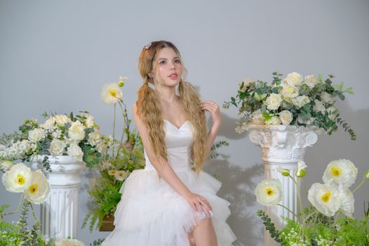 A bride in a white dress sits elegantly with floral arrangements, creating a romantic, indoor scene.