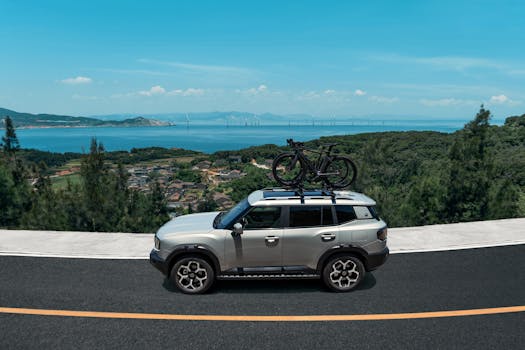 Silver SUV with bicycles on the roof rack driving along a coastal road surrounded by lush green hills and ocean view.