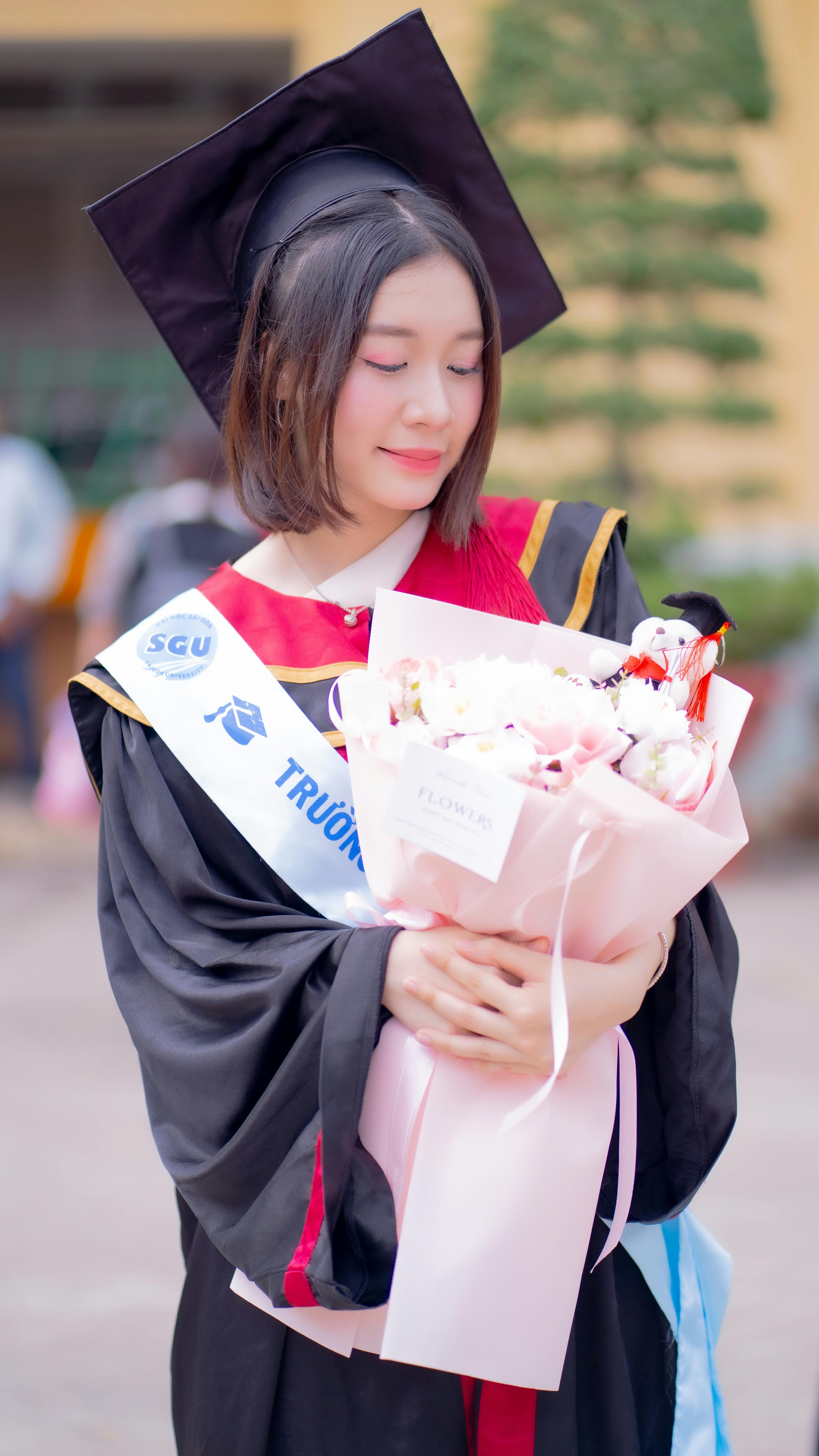 Young graduate in cap and gown holding bouquet, celebrating graduation.