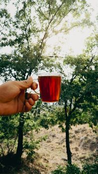 A person holds a glass of tea outdoors with a scenic forest backdrop, capturing nature's tranquility.