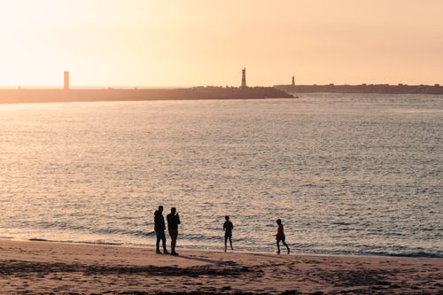 Free Silhouettes of family enjoying a sunset beach scene with a distant lighthouse. Stock Photo